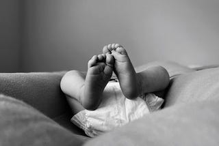 An infant wearing a diaper lying on a cushion with its feet in the air, as seen from below, where only the legs and diaper are visible