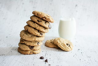 A tower of chocolate chip cookies leans towards a glass of milk and couple more delicious cookies.