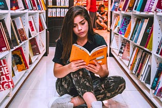 color photpgraph of a young woman reading a book with an orange cover with sitting cross-legged on the floor between two full shelves of books in the library
