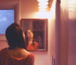 A young woman wearing an orange shirt stands in front of a bathroom mirror, her hair wet from a shower. The reflection is blurry and she is gazing upwards past the top right corner of the frame, conveying that she is contemplating something.