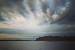 Dramatic clouds above a calm bay, with forested land on the horizon.