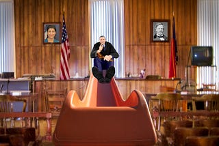 A stern robed judge at the top of an orange slide, holding a gavel, his feet facing us. He is in a courtroom. On the walls behind him hang portraits of Lina Khan and John Sherman. Image: Harrison Keely (modified) https://commons.wikimedia.org/wiki/File:The_Clay_County_Courthouse_courtroom_in_2007.jpg CC BY 4.0 https://creativecommons.org/licenses/by/4.0/deed.en — DocteurCosmos (modified) https://commons.wikimedia.org/wiki/File:Playground_slide_close-up.jpg CC BY 3.0 https://creativecommons