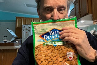 A man holding a bag of Blue Diamond Whole Natural Almonds in what appears to be a kitchen. The package has a green border and displays “Irresistible Snacking!” at the top. The almonds are visible through the packaging, and there’s an American Heart Association certification badge on the packet. The package also mentions “3g Net Carbs” on it. The person is holding the bag up near their face in what seems to be a home kitchen with wooden cabinets visible in the background.
