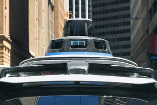 A self-driving car equipped with rooftop sensors and cameras navigates through an urban street canyon, flanked by high-rise buildings.