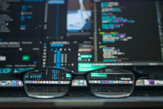 Close-up of eyeglasses focused on the foreground with blurry computer screens showing programming code in the background.