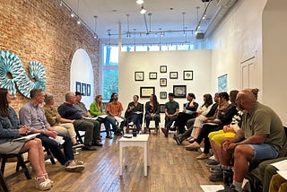A group of 30 people sits in a circle in a room with a one red brick and two white walls. They are engaged in conversation. A white table with a tape recorder rests at the center. On the walls are various works of art.