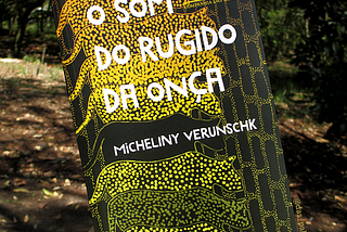 Livro O som do rugido da onça segurado por minha mão, um pouco inclinado para a esquerda, contra o fundo de uma paisagem natural. A capa é preta com desenhos repetidos de uma onça em uma graduação de tons de amarelo a laranja como carimbos de cima a baixo. O título e nome da autora estão grafados em branco.