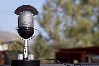 Photo shows a radio microphone in the foreground with a sign over it that reads “ON THE AIR.” The background has blurred a tree.