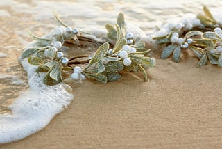 A delicate wreath made of glittering plastic leaves and white pearls rests on beach sand, partially covered by ocean foam. The scene evokes a serene, aesthetic feel.