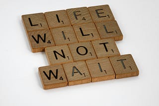Scrabble tiles arranged to spell out “life will not wait”.