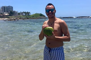The author is shirtless and wearing sunglasses, a white swim short with blue stripes and a neckless. He is holding a coconut while looking at the camera and smiling. He is at the beach and the water is crystal clear blue water. All the way in the back, you see a green hill and to the left, a hotel. The sky is clear as there are no clouds.