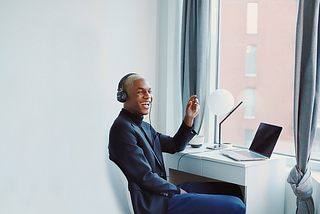 A young, professional Black man at his home desk, wearing headphones and smiling and talking with a laptop open as if on a web call.