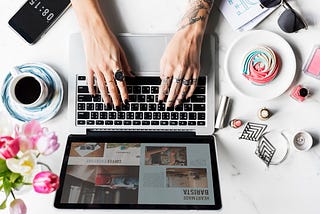 Laptop on a desk with a woman’s hands typing, surrounded by flowers, coffee and personal items