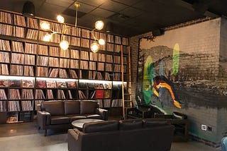 Interior photograph of a lounge with two brown leather couches on either side of a low circular table. The far side wall of the lounge is a floor-to-ceiling bookshelf filled with hundreds of vinyl records and including a wooden ladder attachment. The right-side wall of the space is a painted brick wall.