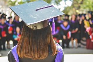 Photo of a person wearing a graduation cap taken from behind, facing a blurry crowd of people in graduating gowns.