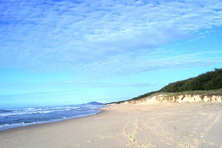 A long view of an empty sandy beach with footprints, the ocean, and a cloudy but magical brilliant blue sky.