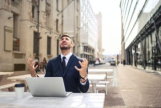 Pria menggunakan setelan formal dengan raut wajah frustrasi sembari melihat ke langit dengan laptop di hadapannya. mengilustrasikan seseorang yang masih kesulitan mendapat pekerjaan karena sering membuat kesalahan paling umum saat membuat CV.