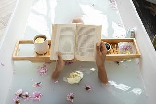 A lady soaking in a bathtub filled with flowers, reading a book. She has a tray set up in the bathtub with holds a cup of her favorite beverage.