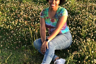 A black woman w/ an afro wearing a tie-dye t-shirt,blue jeans, and white sneakers sitting in a field.