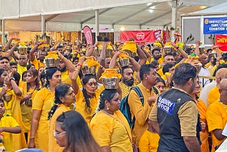Large crowd of people wearing saffron colored clothing, some are carrying silver pots topped with yellow cloth, on their heads.