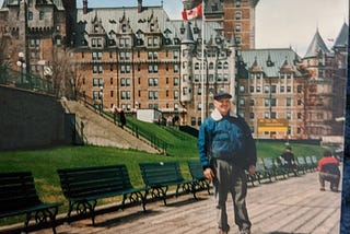 A man in beige pants with a dark blue jacket and a fisherman’s cap poses on a wooden walkway with Château Frontenac — and a Canadian flag waving in the background.