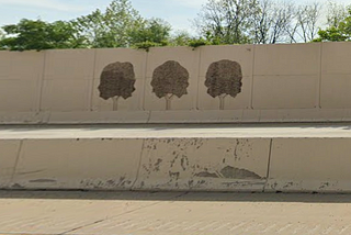 Concrete sound barrier on the side of a highway with three trees painted on it. You can also see a few trees growing behind the barrier that are somewhat taller than it is.