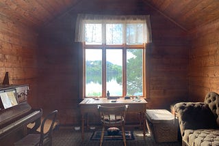A small room with a piano and a desk with a stunning lake and nature view.
