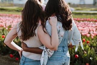 Two female friends with arms wrapped around each other looking out at a field of flowers