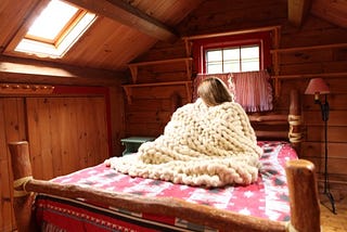 Woman sitting up in bed wrapped in a hand knit bulky blanket.