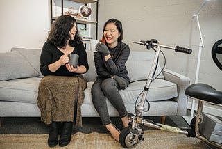 Two disabled women chat on a couch, both holding coffee mugs. An electric lightweight mobility scooter rests nearby.