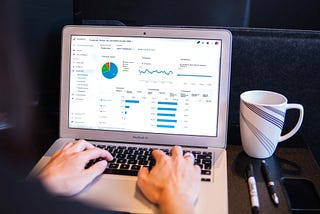 The analytical tool page is on the laptop screen. Two hands, one coffee mug, and two pencils are visible. Someone is checking the SEO performance of their website. Image: https://unsplash.com/