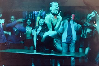 Quinni dancing on top of a table surrounded by drunken partygoers cheering her on. She is knelt down and sticking her tongue out with her fingers in a “V” formation just below. Her outfit is all black with a silk slip dress, a fishnet top underneath, fishnet tights, and black leather combat boots. Her red hair is slicked back and she looks exuberant.