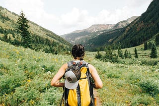 This picture shows a hiker with a yellow backpack walking on a trail with pine trees close by and mountains in the distance.