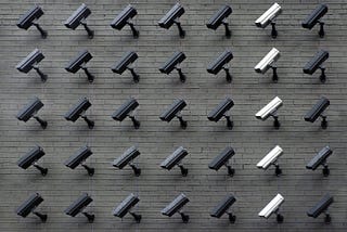 Many CCTV cameras that are differing shades of gray, mounted on a wall