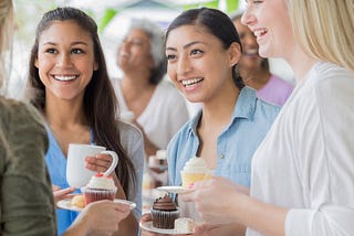 4 women standing talking and smiling each holding a white coffee mug and white plate with a muffin.