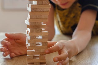 A boy removing a wooden block from a tower that is about to fall over.