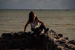 A autora está sentada no final de um pier de pedras, já próxima ao mar, olhando contemplativa para o céu.