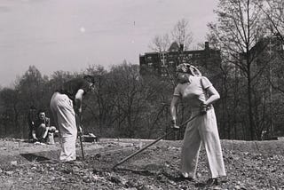 Black-and-white photo of two people tilling bare soil, preparing a garden plot. An apartment building is in the background.