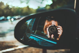 A side mirror of a car, reflecting the image of a woman aiming a digital camera towards the scenery on the side of the road.
