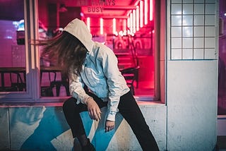 A woman in a hoodie, sitting a a business’s open window, leans away from view. A sign over her head says, “Always busy.”