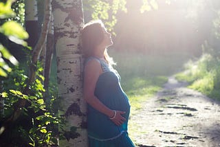 Pregnant woman leaning against tree in sunshine.