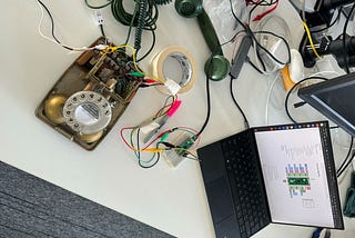 Partially dismantled rotary phone, sharpie, masking tape, rasperry pi pico connected to the some terminals within the phone, and a laptop plugged into the pico strewn across a desk