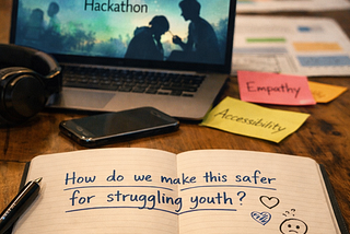 Laptop displays “Building Safer AI for Youth Mental Health Hackathon,” notebook reads “How do we make this safer for struggling youth?”