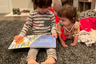 Two kids on a rug. The older child is reading to the younger child.