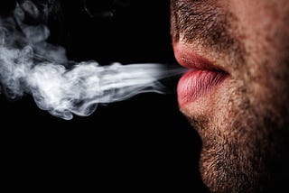 A bearded man in profile blowing out cigarette smoke, showing only his mouth and chin.