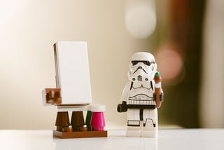 A Stormtrooper Lego figure holding a brush. The figure is standing in front of an easel and a blank canvas surrounded by paint.