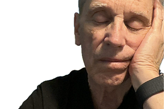 Older man with gray hair wearing a black shirt and smart wristband, resting his head on his hand with eyes closed, appearing to be asleep or deep in thought.