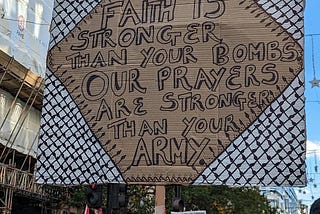 Photo of a placard from a protest with text surrounded by Palestinian keffiyeh material reading ‘Our faith is stronger than your bombs. Our prayers are stronger than your army’.