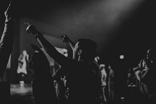 Black and white photo of a young person pointing at a band on a stage with a light overhead coming in from the heavens above.