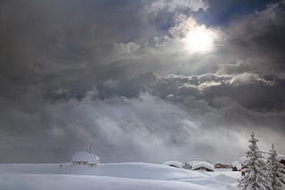 The sun poking through beautiful snow clouds, revealing a landscape in the swiss alps with snow covered huts and fir trees. Very peaceful and giving a sense of a world beyond life.